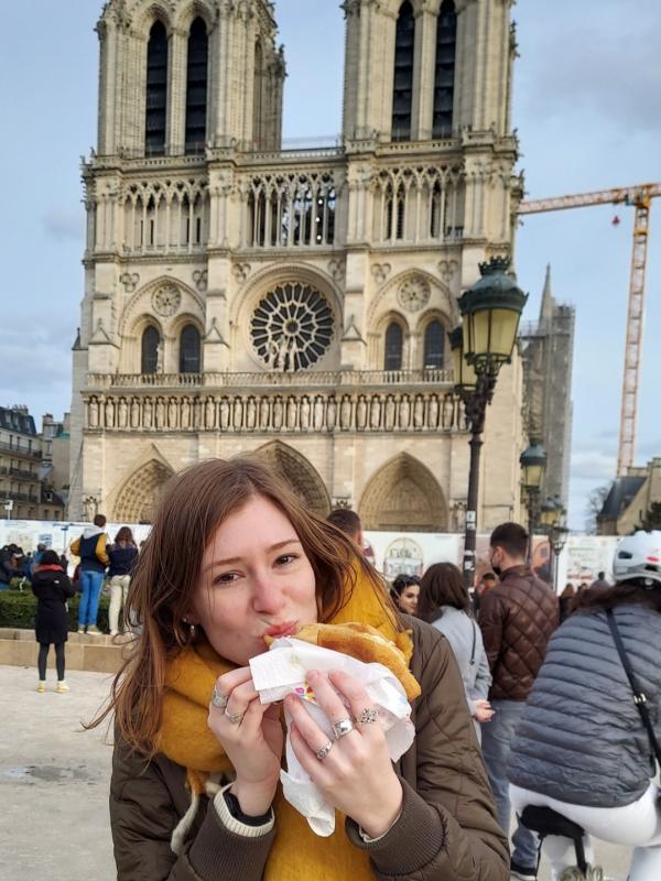 A woman with long brown hair is taking a bite out of bread in front of a gothic cathedral